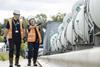 A man and woman wearing orange high vis walk past a pipe