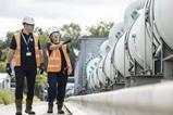 A man and woman wearing orange high vis walk past a pipe