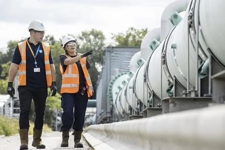 A man and woman wearing orange high vis walk past a pipe